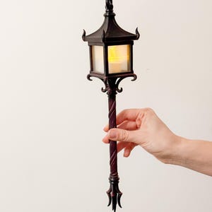 Op de afbeelding: Een decoratieve, donkerbruine lantaarn met een pagode-achtige bovenkant en een warm, geel licht van binnen. De lantaarn wordt vastgehouden door een lange, gedraaide steel met decoratieve accenten. De lantaarn is ongeveer 30 cm hoog.