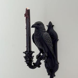 May include: A matte black wall-mounted candle holder featuring a sculpted raven perched on a decorative bracket. The raven holds a single black taper candle in a floral-shaped cup, with dark red wax drips running down the side of the candle.