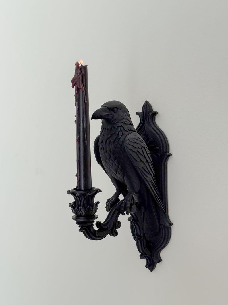 May include: A matte black wall-mounted candle holder featuring a sculpted raven perched on a decorative bracket. The raven holds a single black taper candle in a floral-shaped cup, with dark red wax drips running down the side of the candle.