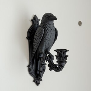 May include: A matte black wall-mounted candle holder featuring a sculpted raven perched on a decorative bracket. The bird is depicted with detailed feathers and a sharp beak, extending a curved arm that supports a floral-shaped candle cup.