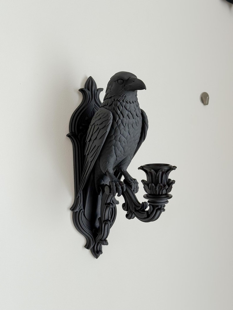 May include: A matte black wall-mounted candle holder featuring a sculpted raven perched on a decorative bracket. The bird is depicted with detailed feathers and a sharp beak, extending a curved arm that supports a floral-shaped candle cup.
