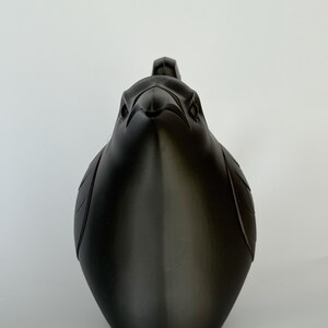 May include: A matte black 3D printed sculpture of a bird, specifically a raven or crow, viewed from the front. The object features stylized, geometric feather details on the wings and a sharp, pointed beak, with a smooth, uniform dark finish.
