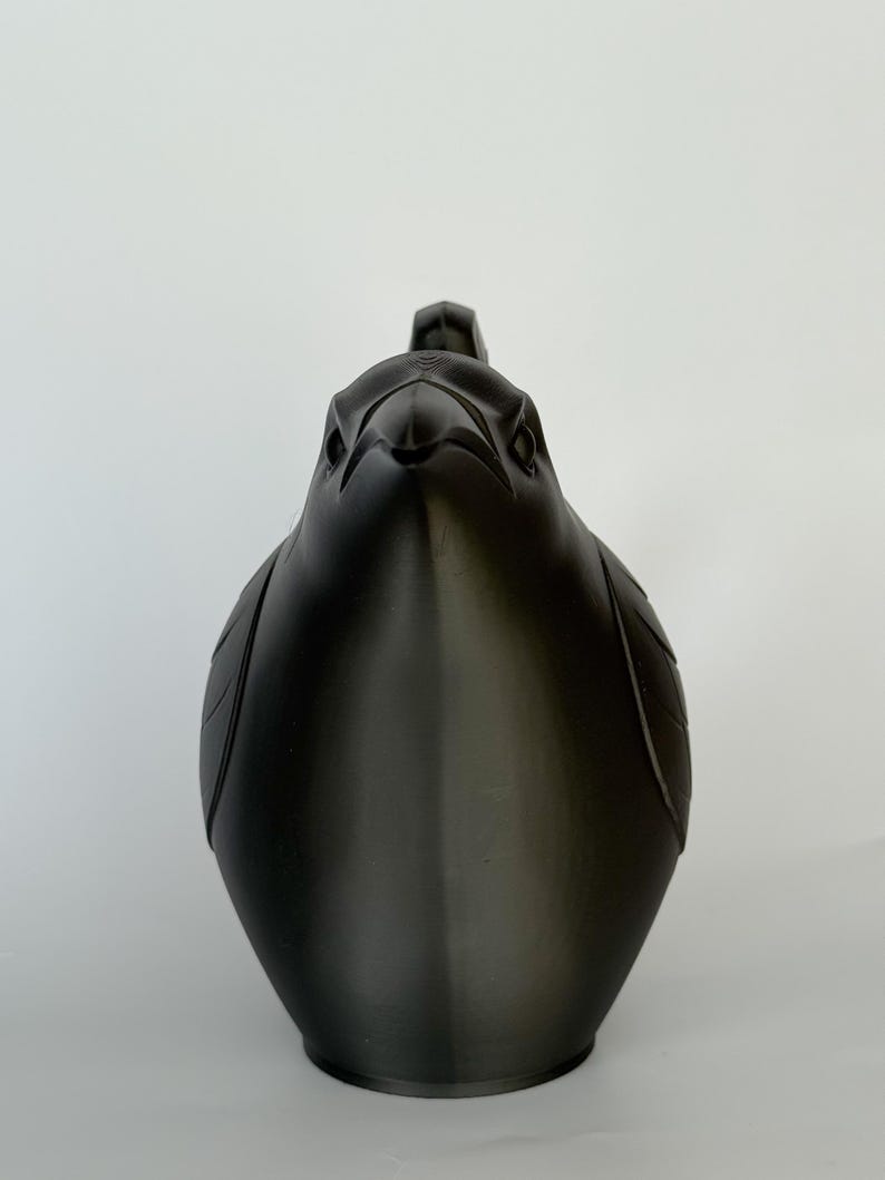 May include: A matte black 3D printed sculpture of a bird, specifically a raven or crow, viewed from the front. The object features stylized, geometric feather details on the wings and a sharp, pointed beak, with a smooth, uniform dark finish.