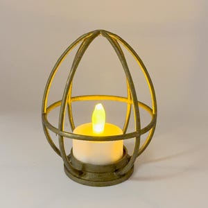 Könnte beinhalten: Ein goldfarbener Metallkerzenhalter mit einem käfigartigen Design. Im Inneren des Käfigs befindet sich eine weiße Kerze mit einer flackernden gelben Flamme. Der Kerzenhalter hat einen runden Sockel und gebogene Stäbe, die eine ovale Form bilden.