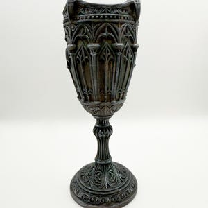 May include: A detailed, dark bronze chalice with gothic architectural patterns. The chalice has a wide, fluted rim, a detailed stem, and a round, decorated base. The design suggests a vintage or antique style, with a height of approximately 20 cm.