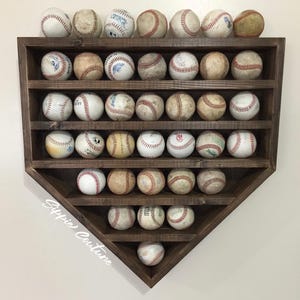 May include: A dark wooden home plate-shaped display case filled with baseballs. The baseballs are white with red stitching, some showing wear. The case has multiple shelves and is mounted on a white wall. The case is designed to hold baseballs.