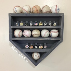May include: A gray baseball-shaped display shelf holding several baseballs and small decorative bottles. The shelf has three levels and is mounted on a light-colored wall. The text "Sippie Couture" is written on the shelf.