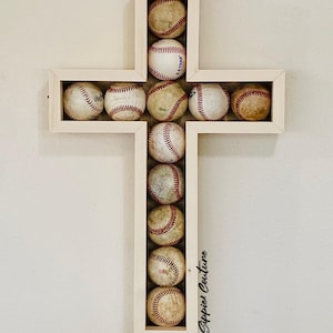 May include: A wooden cross-shaped wall hanging filled with baseballs. The baseballs are arranged within a light-colored wooden frame. The baseballs are white with red stitching and show signs of wear.