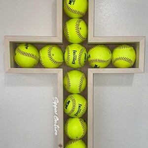 May include: A wooden cross-shaped shelf filled with bright yellow softballs. The softballs are arranged in a cross shape within the wooden frame. The softballs have black stitching and the text "NCAA" and "Fastpitch".