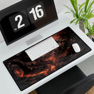 May include: A long, rectangular mouse mat with a dramatic landscape design in fiery reds, oranges, and blacks. A white keyboard and mouse sit on the mat, next to a computer monitor displaying the time. The desk is white, and a plant is in the background.
