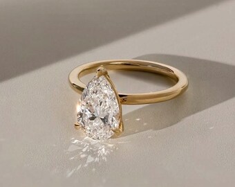 May include: A gold ring with a pear-shaped diamond. The diamond sparkles, reflecting light. The ring is set on a neutral surface, with shadows adding depth. A classic piece of jewelry.