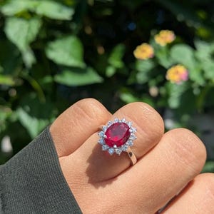 Puede incluir: Un anillo de plata con una gran gema roja oscura de corte ovalado, rodeada de gemas claras más pequeñas. El anillo se presenta sobre un fondo borroso de follaje verde y flores amarillas.