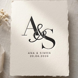 May include: A cream-colored wedding invitation with black text. The design features the initials "A&S" in a decorative font, with the names "ANA & SIMON" and the date "20.06.2026" below. The paper has a textured, deckled edge.