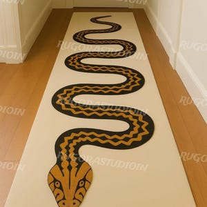 Op de afbeelding: Een beige rechthoekig tapijt met een zwart en goud slangenmotief. De slang kronkelt over de lengte van het tapijt, met een gedetailleerde kop aan de onderkant. Het tapijt ligt in een gang met houten vloer.