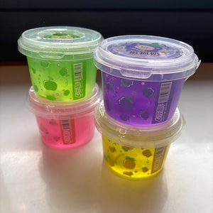 Op de afbeelding: Vier kleine plastic containers gestapeld, elk gevuld met kleurrijke, doorschijnende slijm. De kleuren zijn groen, roze, paars en geel. Elke container heeft een doorzichtige deksel en een etiket met fruitillustraties.