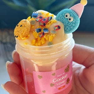 May include: A clear plastic jar filled with pink slime, topped with yellow slime and decorative elements. The decorations include a blue Oreo biscuit with a pink party hat, small car-shaped pieces, and cookie-shaped sprinkles. The jar has a pink label that reads "Cookie Lover."