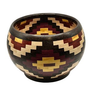 Può includere: Una ciotola in legno fatta a mano con un motivo geometrico. La ciotola presenta un bordo e una base marrone scuro, con un design a mosaico di legno chiaro, bordeaux e giallo. La ciotola è rotonda e ha un'ampia apertura.