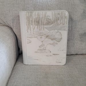May include: White binder with a forest scene and a cartoon fox-like creature. The binder has a stitched edge and a spine. The creature is running on a rock. The word "Ultra Pro" is printed on the bottom.