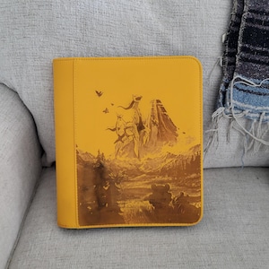 May include: A yellow binder featuring a detailed landscape illustration with figures and mountains. The artwork is in shades of brown and tan, with a few small flying figures. The binder is closed and has a smooth texture.