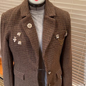 May include: Brown and black houndstooth blazer with decorative brooches. The blazer is displayed on a dress form and features a classic notched lapel, two front pockets, and decorative silver buttons. The brooches include floral and geometric designs.