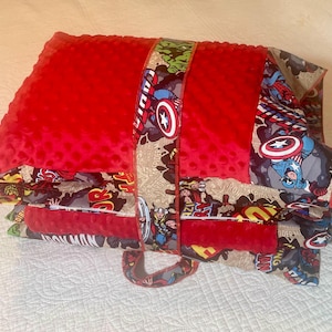 May include: A red, plush, rectangular pillow with a patterned strap featuring comic book characters. The pillow is folded and secured with a strap, showcasing images of Captain America and other heroes. The overall theme is a superhero-themed accessory.