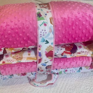 May include: A stack of pink, textured blankets and pillows with a white strap featuring a floral and character design. The blankets and pillows have a princess theme, with images of various princesses.