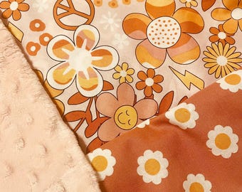 May include: A close-up of a baby blanket with a floral pattern in shades of orange, white, and peach. The design includes daisies, a peace sign, and a smiling flower. A soft, peach-colored, textured fabric borders the floral pattern.