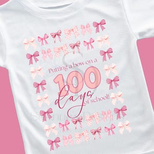Könnte beinhalten: Weißes T-Shirt mit einem rosa Schleifendesign und dem Text "Putting a bow on a 100 days of school!" in Rosa. Das Design umfasst verschiedene rosa Schleifen.