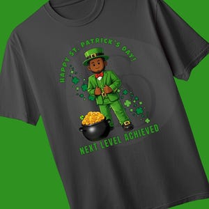 May include: Dark gray t-shirt featuring a cartoon illustration for St. Patrick's Day. The design includes a person in a green suit and hat, a pot of gold, and the text "Happy St. Patrick's Day!" and "Next Level Achieved."