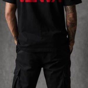 May include: Black t-shirt with the word "VANTA" in red block letters across the back. The shirt is paired with black cargo trousers and white trainers. The person is standing in front of a grey background.
