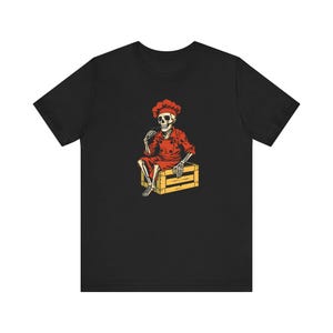 Könnte beinhalten: Schwarzes T-Shirt mit einem Cartoon-Skelettkoch-Design. Das Skelett, in roter Kochmütze und Uniform, sitzt auf einer gelben Holzkiste und hält ein Utensil. Die Illustration hat einen Vintage-Stil, mit Fokus auf das Kochthema.