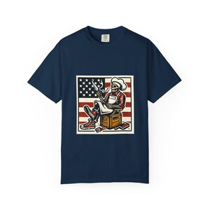 Könnte beinhalten: Marineblaues T-Shirt mit einem quadratischen Motiv eines Skelettkochs mit einem Messer vor einer amerikanischen Flagge. Das Skelett trägt eine Kochmütze, eine Schürze und rote Turnschuhe. Eine Holzkiste, eine Gabel und ein Messer sind ebenfalls abgebildet.