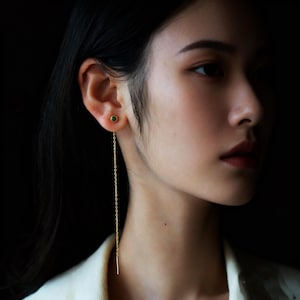 Puede incluir: Primer plano de una persona con pendientes dorados. Cada pendiente presenta un pequeño y redondo perno de piedra verde y una larga y delicada cadena dorada. El fondo oscuro resalta las joyas.