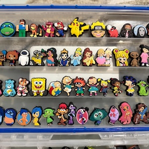 Puede incluir: Un contenedor de almacenamiento de plástico transparente lleno de dijes de zapatos coloridos. Los dijes presentan varios personajes de dibujos animados, incluidos las Tortugas Ninja, Bob Esponja y personajes de películas de Disney.