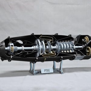 May include: A detailed cross-section model of a PT6E turboprop engine, showcasing internal components. The engine is black with silver and gold-colored gears and blades. The model is mounted on a gray stand, with the text "PT6E" visible on a small plate.