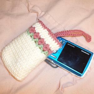 May include: A cream-colored, crocheted camera case with a pink and green floral design. The case has a matching pink strap and holds a blue Olympus digital camera. The case is handmade and appears to be soft and protective.