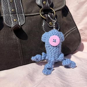 May include: A light blue crocheted creature keyring with a pink button detail. The keyring is attached to a brown suede handbag with leather straps. The background is a pink fabric.
