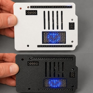 May include: A rectangular electronic circuit board enclosure featuring a white or black textured finish, multiple pin headers, vertical ventilation slots, and a central square mesh cutout revealing internal blue LED lights.