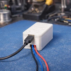 Pode incluir: Um dispositivo eletrônico retangular branco com uma porta USB-C e três fios coloridos. O dispositivo está conectado a um cabo preto. O fundo é uma superfície azul com ferramentas.