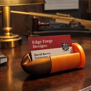 Könnte beinhalten: Ein orange-brauner Visitenkartenhalter in Form einer Patrone. Er enthält eine Visitenkarte mit dem Text "Edge Forge Designs" und "David Reeves, Businessman, AL". Er steht auf einem Holzschreibtisch, mit einer Lampe und einem Notizbuch im Hintergrund.
