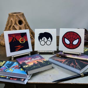 May include: Three small embroidered canvases on wooden easels. One features a volcano scene, another Harry Potter's silhouette, and the third, Spider-Man's mask. Books and a pen are also visible.