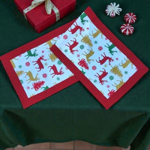 May include: Two rectangular Christmas placemats with a red border. The center features a white background with colorful reindeer, snowflakes, and Christmas trees. A red gift box with a white ribbon is in the background.