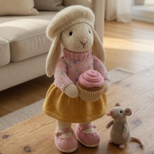 May include: A handmade knitted bunny doll wearing a pink sweater, yellow skirt, and beret, holding a cupcake. A small knitted mouse is beside the bunny on a wooden table. The scene is set in a cozy living room with a beige sofa and natural light.