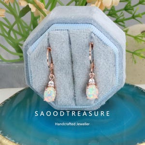 May include: Rose gold earrings with oval opal and clear stone accents, displayed in a light blue velvet jewelry box. The earrings are on a blue and teal surface. The text "SAOODTREASURE" and "Handcrafted Jeweller" are visible.