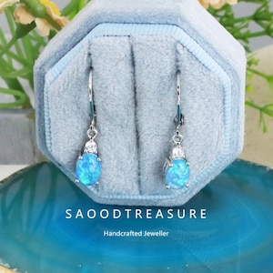 May include: A pair of silver drop earrings with oval, light blue opal stones. Each earring has a small cluster of clear stones above the opal. The earrings are displayed in a light blue velvet jewelry box. The text "SAOODTREASURE" and "Handcrafted Jeweller" are visible.