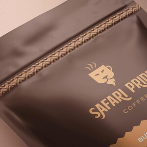 May include: A brown coffee bag with a gold geometric pattern along the top edge. The bag features the words "SAFARI PRIDE COFFEE" in gold, along with a lion head logo. The bag is sealed and ready for use.