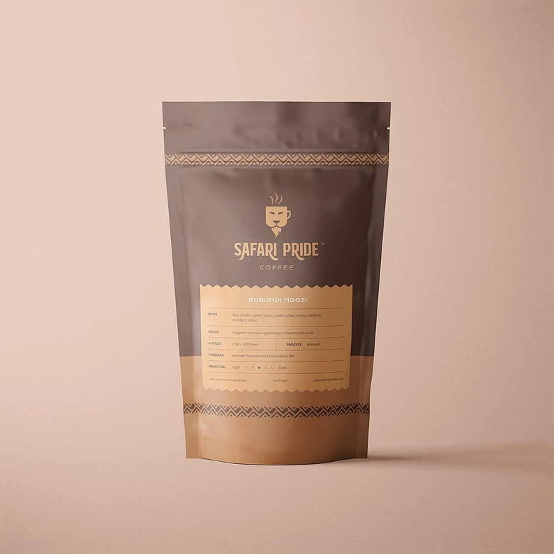 May include: A brown and tan coffee bag with the text "Safari Pride Coffee" and "Burundi Ngozi". The bag has a decorative pattern at the top and bottom. The bag is standing upright against a light pink background.