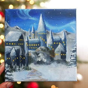 Puede incluir: Pintura en lienzo cuadrada de un castillo bajo un cielo estrellado. El castillo tiene múltiples torres y está pintado en tonos grises y azules. El cielo presenta un efecto de aurora boreal azul. La pintura está ambientada en un paisaje nevado.