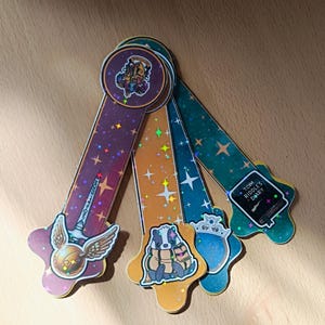 May include: Four decorative bookmarks with different designs. Each bookmark has a unique theme, including a golden snitch, a badger, a crown, and a book with the text "Tom Riddle's Diary". The bookmarks are decorated with stars and glitter.