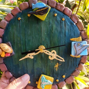 Puede incluir: Un reloj redondo artesanal con un diseño de puerta verde, con un borde de ladrillo marrón. La esfera del reloj tiene detalles dorados y elementos decorativos, como un pez y pan. Las manecillas del reloj son doradas.